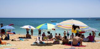 Mehrere Urlauber befinden sich an einem Strand. Die Sonne scheint und einige Sonnenschirme sind aufgespannt. Ein paar Menschen liegen am Strand und sonnen sich. Andere baden im Meer.