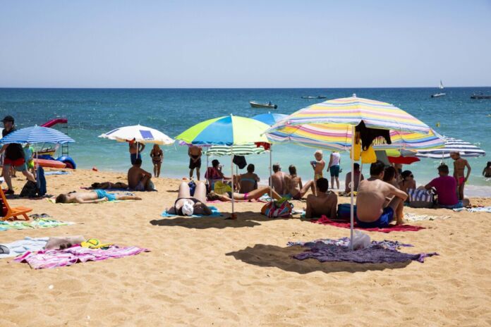 Mehrere Urlauber befinden sich an einem Strand. Die Sonne scheint und einige Sonnenschirme sind aufgespannt. Ein paar Menschen liegen am Strand und sonnen sich. Andere baden im Meer.