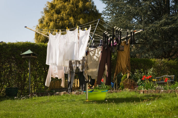 An einer Wäschespinne in einem Garten hängt Wäsche in verschiedenen Farben und Größen. Der Garten ist gepflegt und ist von Bäumen und einer Hecke umgeben.