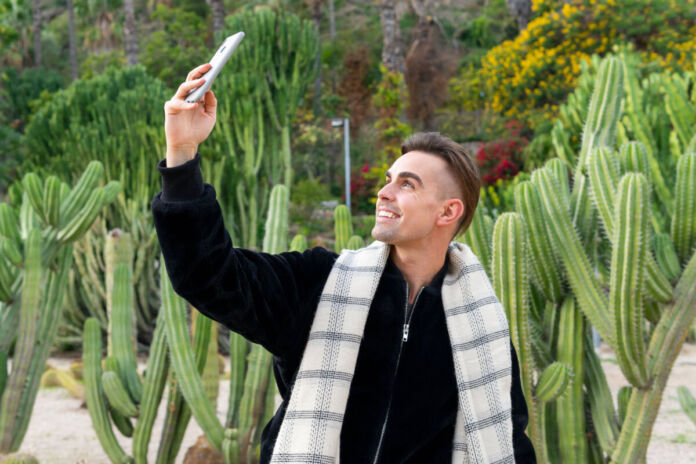 Ein junger Mann in einem stylishen Outfit steht in der Mitte einiger Kakteen und macht von sich und der Umgebung ein Selfie mit dem Smartphone.