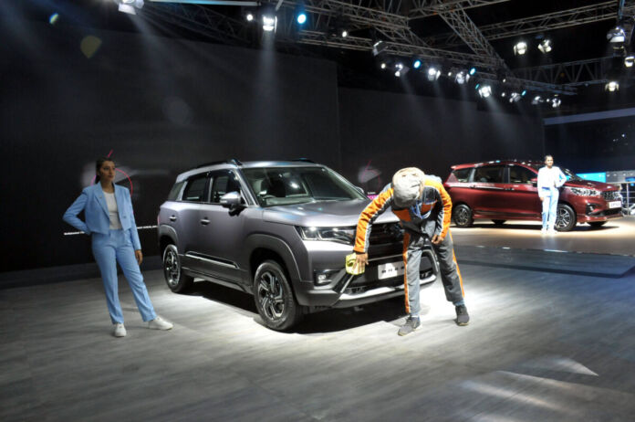 Ein grauer Suzuki Maruti auf einer Ausstellung im hellen Scheinwerferlicht. Die Vorderseite wird von einem Mann geputzt. Eine junge Frau steht daneben.