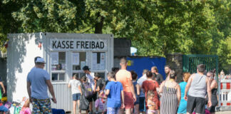 An der Kasse des Freibades stehen viele Besucher Schlange. Hier kaufen sie ihre Eintrittskarte und bezahlen mit ihrem Geld, um später im Schwimmbad zu baden.