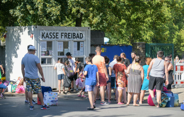 An der Kasse des Freibades stehen viele Besucher Schlange. Hier kaufen sie ihre Eintrittskarte und bezahlen mit ihrem Geld, um später im Schwimmbad zu baden.