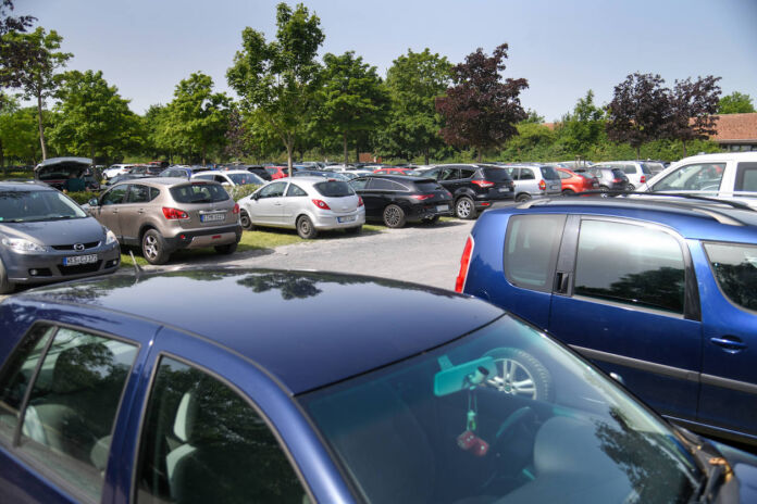 Unzählige Autos stehen an einem heißen Tag nebeneinander auf einem Parkplatz. Die Pkw stehen auf einem Stück Wiese, der Gehweg ist aus Kies. Im Hintergrund stehen Bäume. Die Parkfläche bietet kaum Schatten.