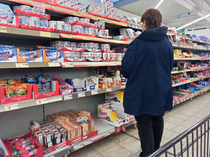 Eine Frau steht in einem Discounter oder Supermarkt vor einem Regal voller Süßigkeiten. Es sind Schokolade und andere Artikel bekannter Marken zu sehen.