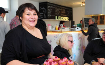 Eine Frau steht glücklich in ihrem Café, in der Hand ein Tablett mit Muffins. Es handelt sich um veganes Gebäck, Kuchen und Torten. Im Hintergrund sitzen Gäste und genießen die Speisen.