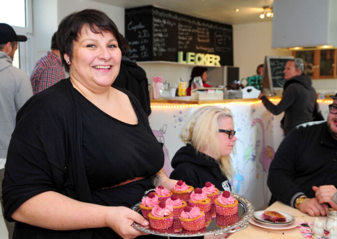 Eine Frau steht glücklich in ihrem Café, in der Hand ein Tablett mit Muffins. Es handelt sich um veganes Gebäck, Kuchen und Torten. Im Hintergrund sitzen Gäste und genießen die Speisen.