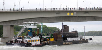 Ein großes Schiff fährt über den Fluss und hat ein U-Boot geladen. Am Rheinufer und auf der Brücke schauen viele interessierte Menschen zu, wie das U-Boot an ihnen vorbeifährt. Es ist auf dem Weg ins Museum.