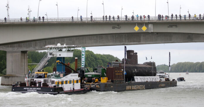 Ein großes Schiff fährt über den Fluss und hat ein U-Boot geladen. Am Rheinufer und auf der Brücke schauen viele interessierte Menschen zu, wie das U-Boot an ihnen vorbeifährt. Es ist auf dem Weg ins Museum.