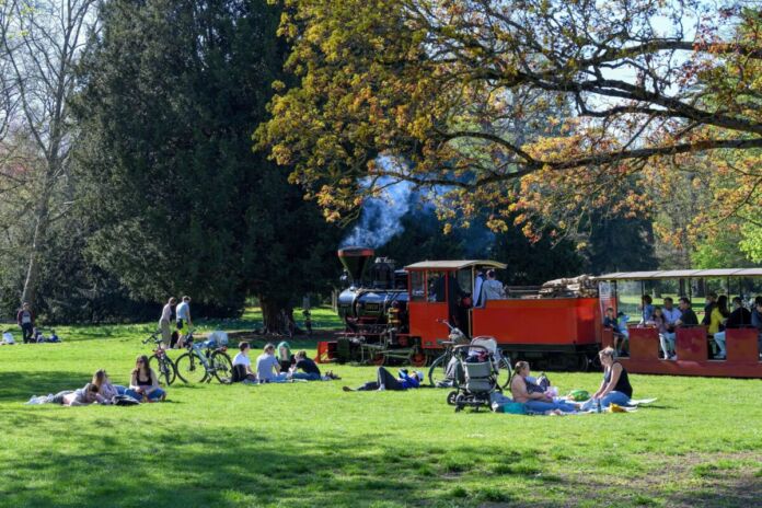 Die Schlossgartenbahn fährt durch den Park vorbei an Bürgern, die es sich gut gehen lassen auf der Wiese. Sie liegen auf einer Decke oder machen ein Picknick.