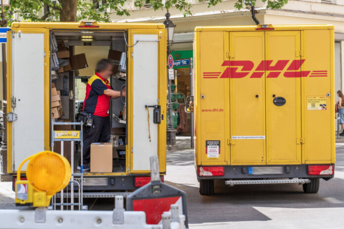 Ein Paketzusteller steht in seinem Fahrzeug von DHL und sortiert Briefe und Pakete. Viele größere und kleinere Pakete sind in Regalen an den Wänden des Fahrzeugs aufgereiht und aufeinander gestapelt. Wegen des Gewichts so manches Pakets bekommen immer mehr DHL-Paketboten Rückenprobleme.