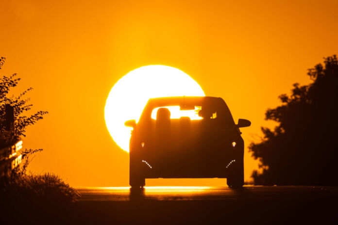 Ein Auto fährt eine Straße entlang. Man sieht nur schwarze Schatten von einem Auto, der Straße und von den Bäumen und Büschen rechts und links. Vor einem orange-gelben Himmel geht gerade die Sonne auf.