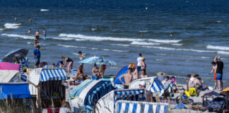 Viele Urlauber sind am Strand, liegen im Sand und sonnen sich. Einige entspannen sich oder baden im Meer. Am Strand stehen auch Strandkörbe.