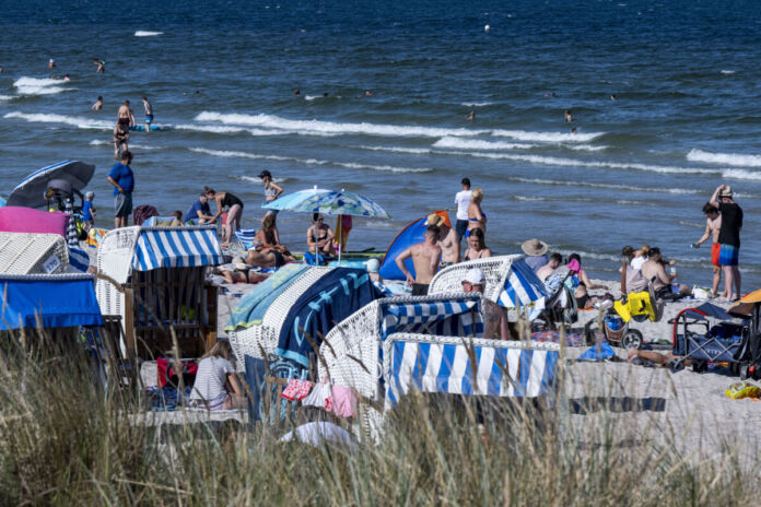 Viele Urlauber sind am Strand, liegen im Sand und sonnen sich. Einige entspannen sich oder baden im Meer. Am Strand stehen auch Strandkörbe.