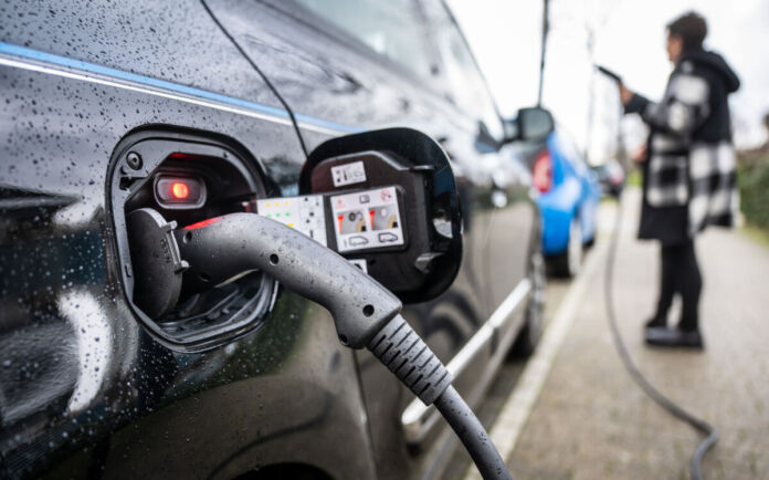 Ein Close-up auf das Ladekabel, das in einem E-Auto steckt. Die Batterie des Elektroautos wird aufgeladen. Auf der Karosserie des Stromers sind vereinzelt Regentropfen, während eine Frau im Hintergrund unscharf steht.