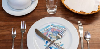 Auf einem Tisch in einem Restaurant steht vorn am Platz ein Teller, auf dem sich unter einem gekreuzten Messer und einer Gabel zwei Zwanzig-Euro-Scheine befinden. Links neben dem Teller liegen zwei Gabeln, rechts ein Messer und ein Löffel. Außerdem steht dort eine umgedrehte Kaffeetasse auf einem Unterteller, ein Glas mit Wasser und ein Brotkorb.