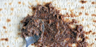 Auf einem Brot, das aussieht wie Matzen, wird mit einem Löffel gerade eine braune Paste verteilt. In dieser sind Stückchen aus Walnüssen zu erkennen.