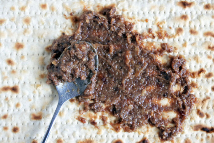 Auf einem Brot, das aussieht wie Matzen, wird mit einem Löffel gerade eine braune Paste verteilt. In dieser sind Stückchen aus Walnüssen zu erkennen.