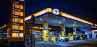 Eine Tankstelle bei Nacht. Die Zapfstelle gehört zu der Tankstellenkette Jet. Die Preistafel im Vordergrund trägt blaue Ziffern und ist gelb beleuchtet. Im Hintergrund tanken zwei silberne Autos.