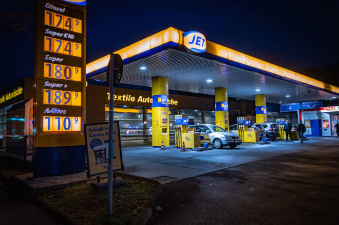 Eine Tankstelle bei Nacht. Die Zapfstelle gehört zu der Tankstellenkette Jet. Die Preistafel im Vordergrund trägt blaue Ziffern und ist gelb beleuchtet. Im Hintergrund tanken zwei silberne Autos.