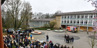 Draußen auf dem Schulhof findet ein Einsatz statt. Im Vordergrund versammeln sich die Schüler, im Hintergrund steht ein Einsatzfahrzeug der Feuerwehr vor dem Schulgebäude.