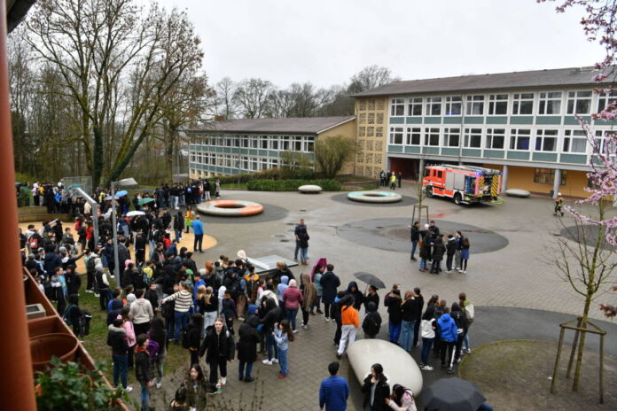 Draußen auf dem Schulhof findet ein Einsatz statt. Im Vordergrund versammeln sich die Schüler, im Hintergrund steht ein Einsatzfahrzeug der Feuerwehr vor dem Schulgebäude.