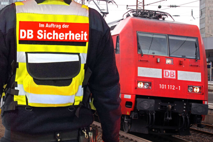 Ein Sicherheitsbeamter steht an einem Bahnhof vor einem roten Zug und überwacht den Bahnhof. Der Mann ist von hinten zu sehen und trägt eine gelbe Warnweste und sorgt für Sicherheit an einem zu gefährlichen Bahnhof.