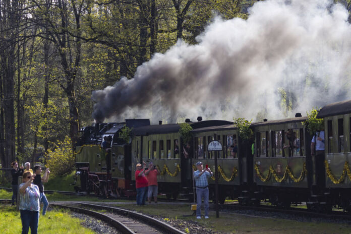 Mehrere Touristen oder Schaulustige stehen am Rand und fotografieren eine nostalgische Eisenbahn. Diese Dampflok fährt auf einer traditionellen Strecke mitten durch die Natur und ist ein echtes Highlight für alle Besucher.