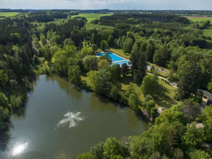 Ein See liegt wunderschön umrahmt von Bäumen und unberührter Natur. Die Luftaufnahme zeigt das Wasser des Sees und die Naturlandschaft. Daneben ist ein Freibad zu sehen. Das Schwimmbad liegt mitten in der Natur.