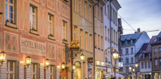 Ein Gasthof befindet sich in einem schönen Gebäude in einer Fußgängerzone. Männer und Frauen laufen an dem Hotel vorbei. Die Straße wird von Laternen beleuchtet.