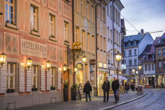 Ein Gasthof befindet sich in einem schönen Gebäude in einer Fußgängerzone. Männer und Frauen laufen an dem Hotel vorbei. Die Straße wird von Laternen beleuchtet.