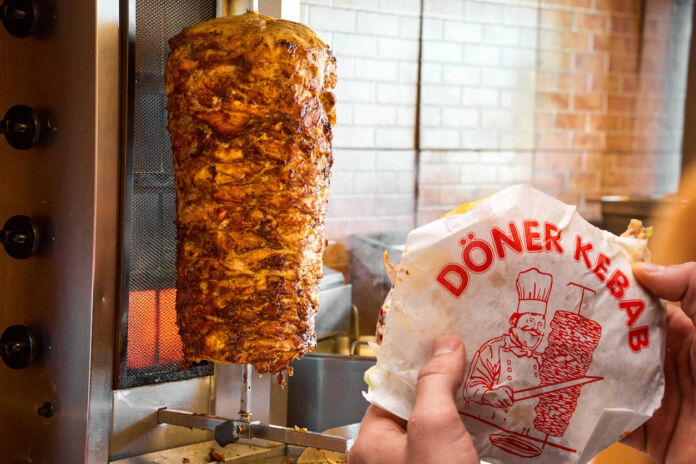 Ein saftiger, mit Fleisch bestückter Dönerspieß dreht sich in einem Dönerladen. Im Vordergrund hält jemand Fladenbrot in typischem Papier für Döner Kebab.