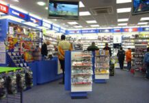 Kunden stehen in einem Laden von GameStop, der Computerspiele und Videospiele zu Konsolen von GameBoy, Playstation und Xbox verkauft. Im Vordergrund hängen Controller, daneben ist ein Aufsteller mit Super Mario.