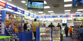 Kunden stehen in einem Laden von GameStop, der Computerspiele und Videospiele zu Konsolen von GameBoy, Playstation und Xbox verkauft. Im Vordergrund hängen Controller, daneben ist ein Aufsteller mit Super Mario.