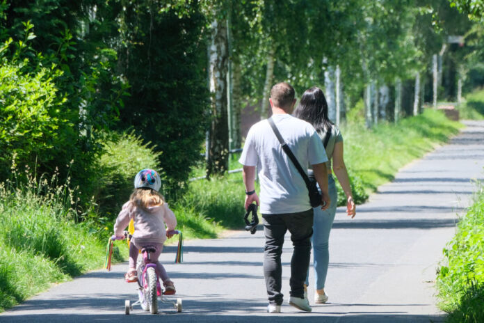 Mutter und Vater gehen gemeinsam mit ihrem Kind spazieren. Das Kind fährt auf einem kleinen Kinderfahrrad mit Stützräder nebenher. Sie verbringen gemeinsame Familienzeit.