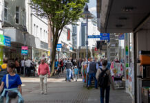 An einem offensichtlichen Tag des Wochenendes bummeln viele Menschen durch die Innenstadt und laufen an den offenen Geschäften vorbei. Einige kommen mit Einkaufstüten aus den Läden. Die Fußgängerzone ist gut besucht.