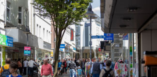 An einem offensichtlichen Tag des Wochenendes bummeln viele Menschen durch die Innenstadt und laufen an den offenen Geschäften vorbei. Einige kommen mit Einkaufstüten aus den Läden. Die Fußgängerzone ist gut besucht.