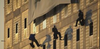Mehrere Klima-Aktivisten klettern die Fassade eines Gebäudes hoch und bringen ein Plakat an. Sie rollen dieses gerade ab und lassen sich an den Seilen langsam herunter.