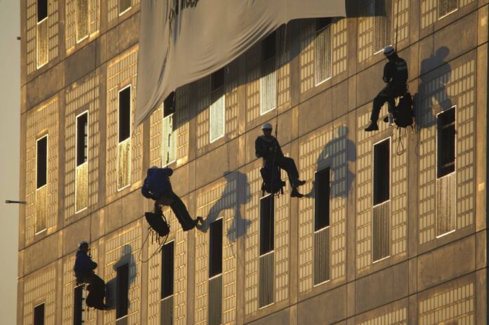 Mehrere Klima-Aktivisten klettern die Fassade eines Gebäudes hoch und bringen ein Plakat an. Sie rollen dieses gerade ab und lassen sich an den Seilen langsam herunter.