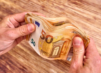 Die beiden Hände eines Mannes halten ein Bündel von 50-Euro-Scheinen. Dabei scheint es sich um mehrere Hundert Euro zu handeln. Die Hände sind auf einem Holztisch abgestützt.
