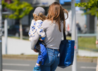 Eine Mutter trägt ihr Kind zur Kindertagesstätte, beziehungsweise zur Kita. Dort wird es über den gesamten Tag betreut, damit die Eltern arbeiten gehen können. Der Junge ist auf dem Arm der Mutter. Sie trägt noch eine Tasche und hält seine Brottasche in der Hand.