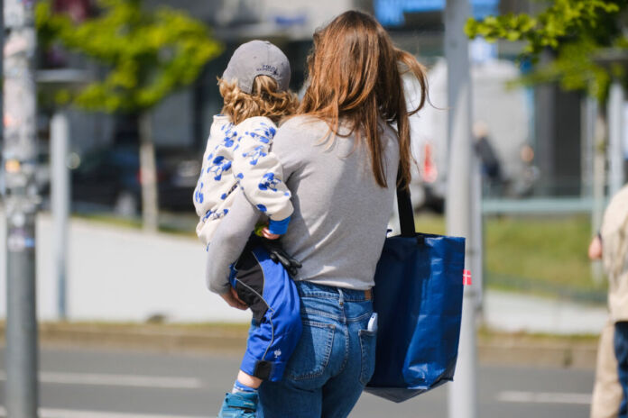 Eine Mutter trägt ihr Kind zur Kindertagesstätte, beziehungsweise zur Kita. Dort wird es über den gesamten Tag betreut, damit die Eltern arbeiten gehen können. Der Junge ist auf dem Arm der Mutter. Sie trägt noch eine Tasche und hält seine Brottasche in der Hand.