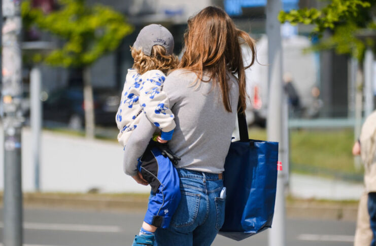 Eine Mutter trägt ihr Kind zur Kindertagesstätte, beziehungsweise zur Kita. Dort wird es über den gesamten Tag betreut, damit die Eltern arbeiten gehen können. Der Junge ist auf dem Arm der Mutter. Sie trägt noch eine Tasche und hält seine Brottasche in der Hand.