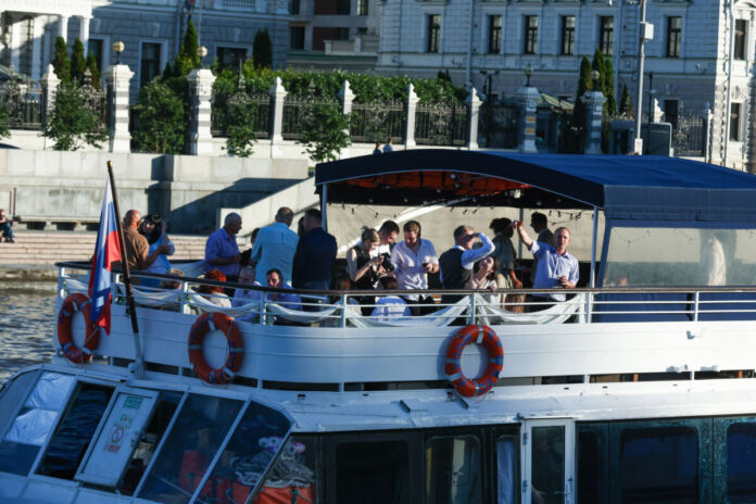 Die Passagiere und Fahrgäste befinden sich an Deck eines großen Fahrgastschiffes, wo sie Getränke und Speisen zu sich nehmen. Das Schiff befindet sich auf einer Rundfahrt auf einem Fluss oder einem anderen Gewässer.