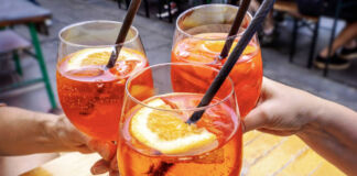 Drei Gäste prosten sich mit Aperol Spritz in großen Weingläsern zu. In jedem Glas befinden sich Prosecco, Aperol, eine Orangenscheibe und Eis. Außerdem haben sie noch einen schwarzen Strohhalm bekommen. Sie sitzen in einem Biergarten auf einer großen Terrasse.