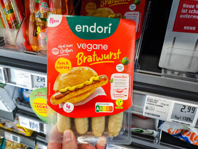 Die Hand von einem Kunden hält vor einem Supermarkt-Regal eine Packung eines Fleischersatzproduktes in einer Hand. Es sind vier vegane Bratwürste von endori verpackt. Eine Supermarktkette will nun weitere Filialen auf vegan umstellen.