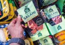 Die Hand eines Kunden greift in das Kühlregal eines Supermarktes oder Discounters und nimmt eine Packung veganer Salami heraus. Neben diesem Produkt befinden sich noch viele weitere vegane Produkte im Kühlregal.