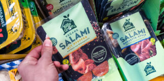 Die Hand eines Kunden greift in das Kühlregal eines Supermarktes oder Discounters und nimmt eine Packung veganer Salami heraus. Neben diesem Produkt befinden sich noch viele weitere vegane Produkte im Kühlregal.