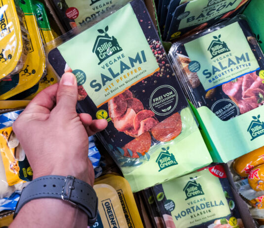 Die Hand eines Kunden greift in das Kühlregal eines Supermarktes oder Discounters und nimmt eine Packung veganer Salami heraus. Neben diesem Produkt befinden sich noch viele weitere vegane Produkte im Kühlregal.