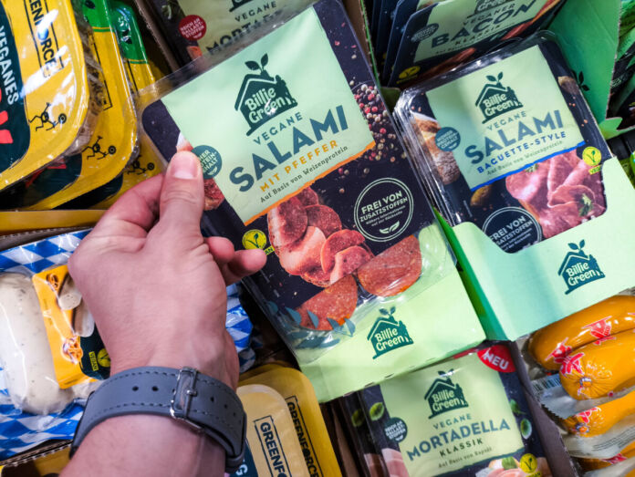 Die Hand eines Kunden greift in das Kühlregal eines Supermarktes oder Discounters und nimmt eine Packung veganer Salami heraus. Neben diesem Produkt befinden sich noch viele weitere vegane Produkte im Kühlregal.